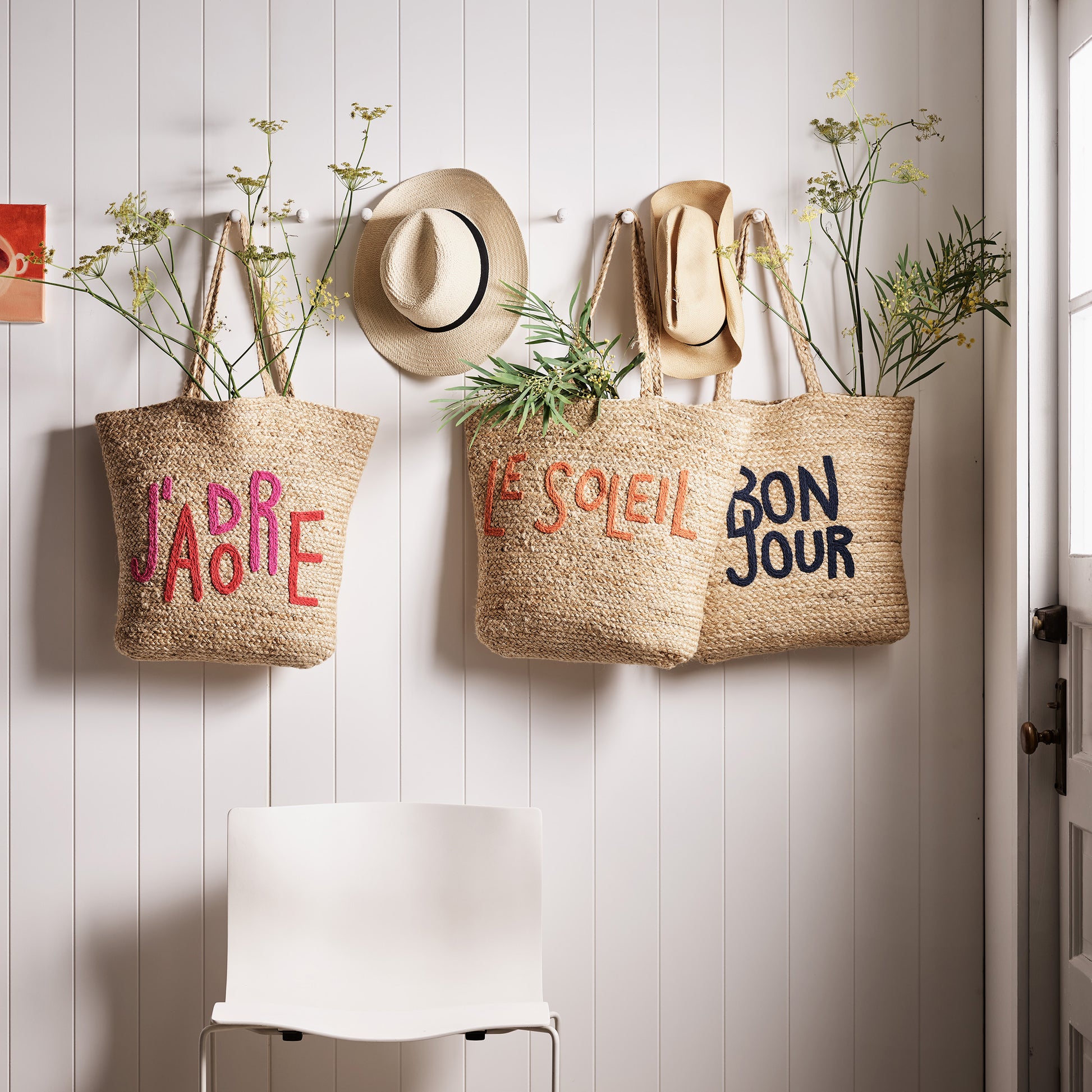 Natural jute woven tote bag with "Bon Jour" embroidered in navy blue, hanging on a white wall.