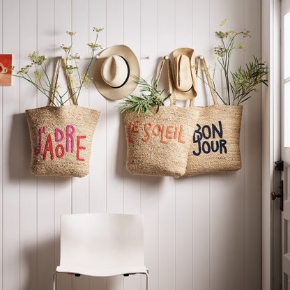 Natural jute woven tote bag with "Bon Jour" embroidered in navy blue, hanging on a white wall.