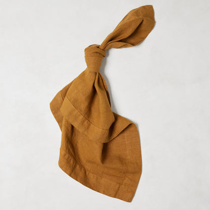 Brass HG Signature hand-dyed linen napkin, 22-inch square, tied in a knot, on a white background.