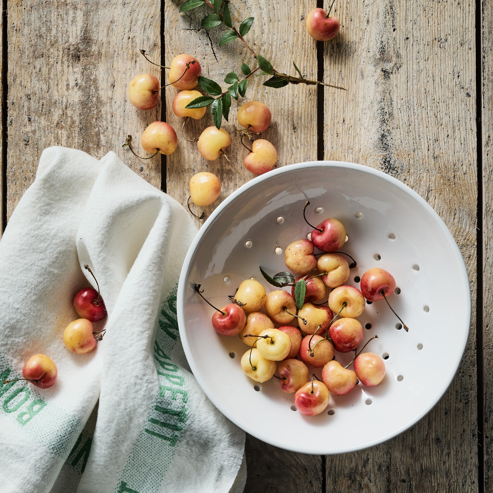 White linen hand towel with green "Bon Appetit" text, next to a white colander filled with Rainier cherries.