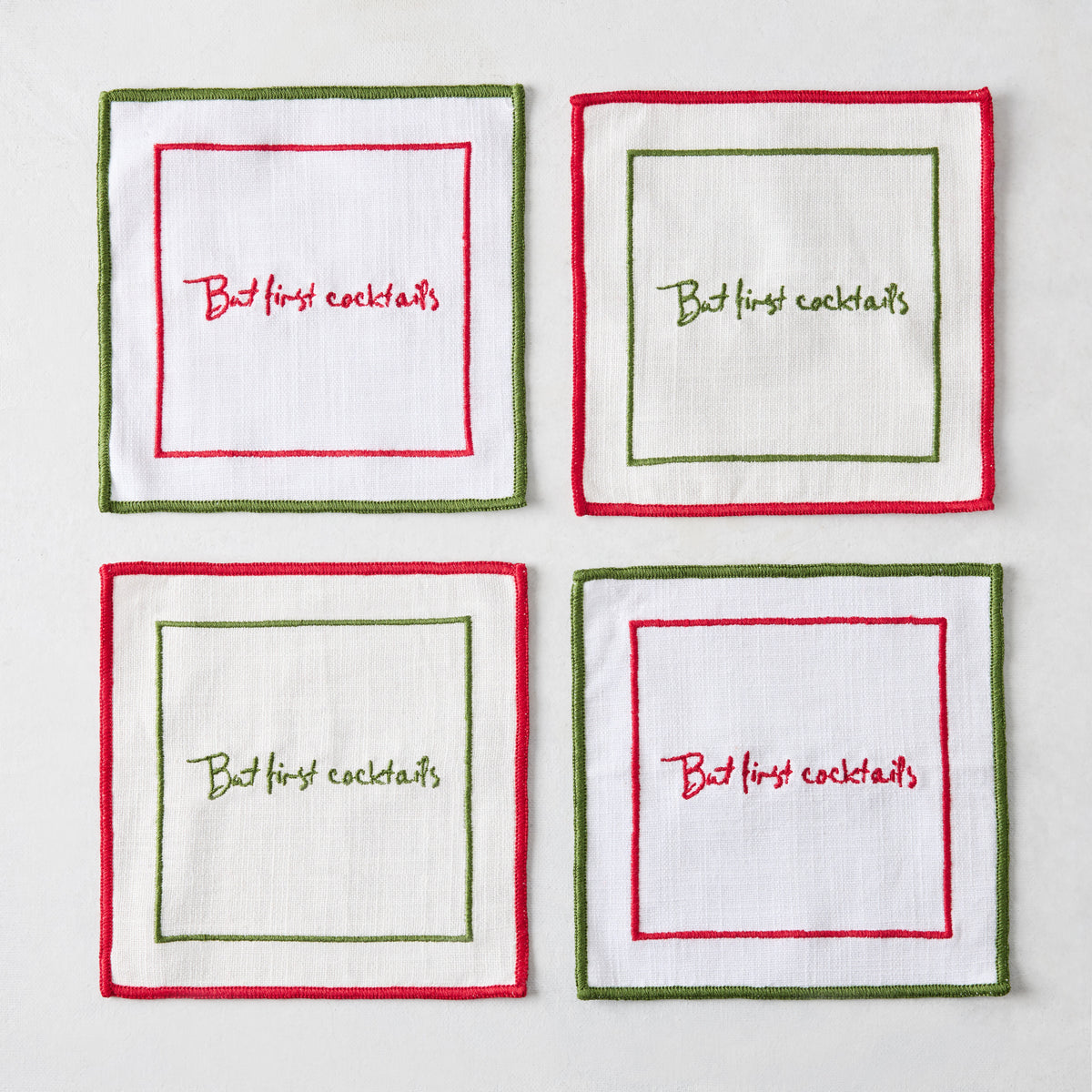 Set of four square white linen coasters with red and green embroidered borders and But First Cocktails text.