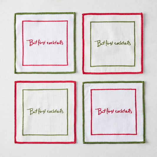 Set of four square white linen coasters with red and green embroidered borders and But First Cocktails text.
