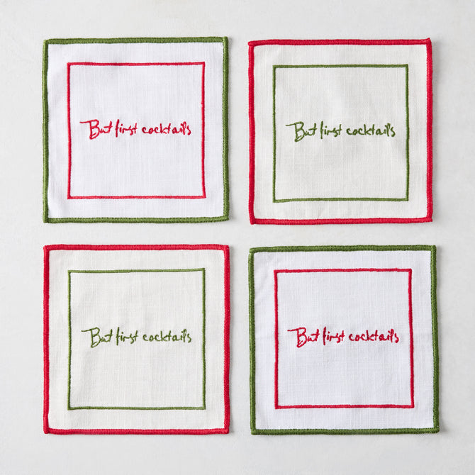 Set of four square white linen coasters with red and green embroidered borders and But First Cocktails text.
