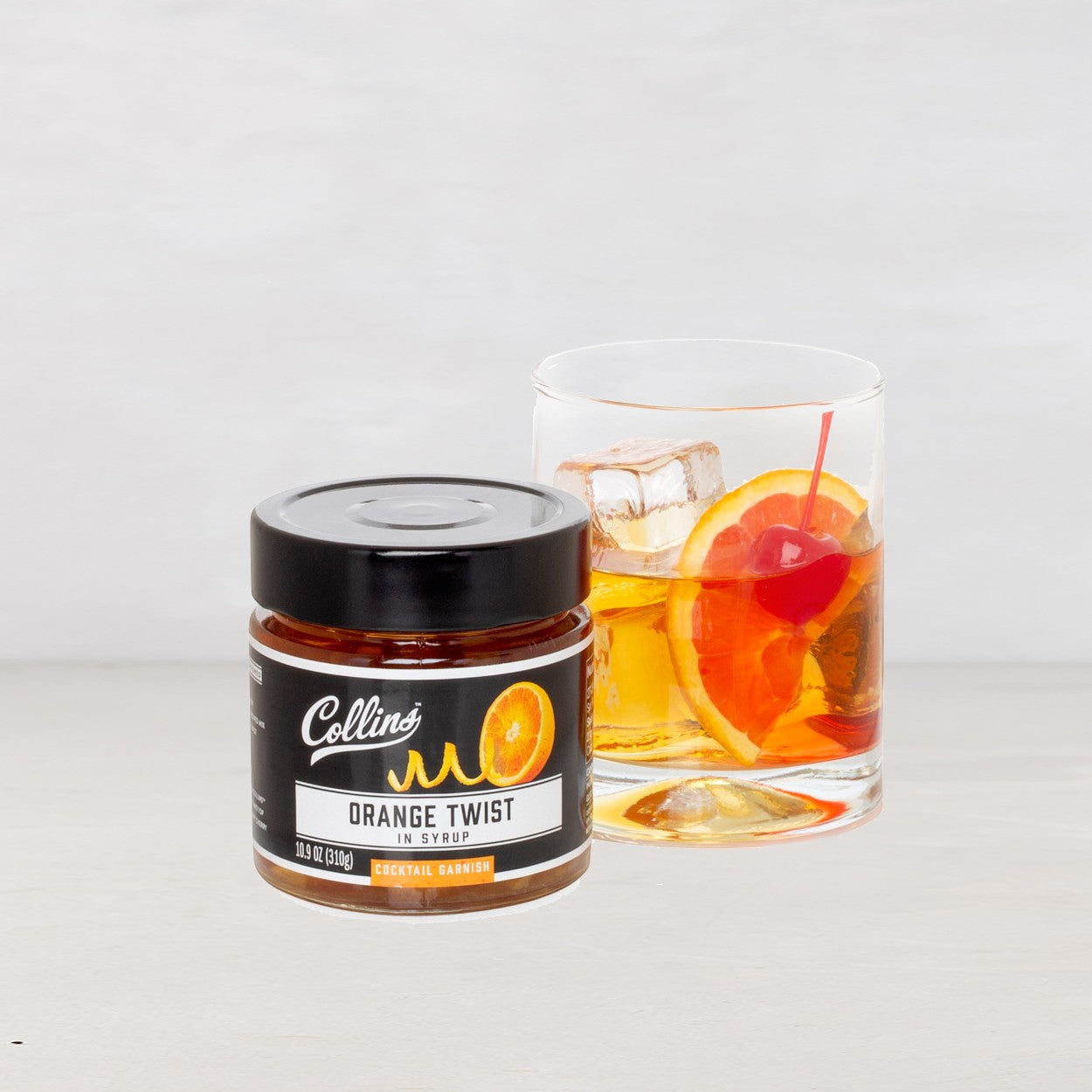 Collins Orange Twist in Syrup cocktail garnish jar next to a whiskey cocktail with an orange slice and cherry.