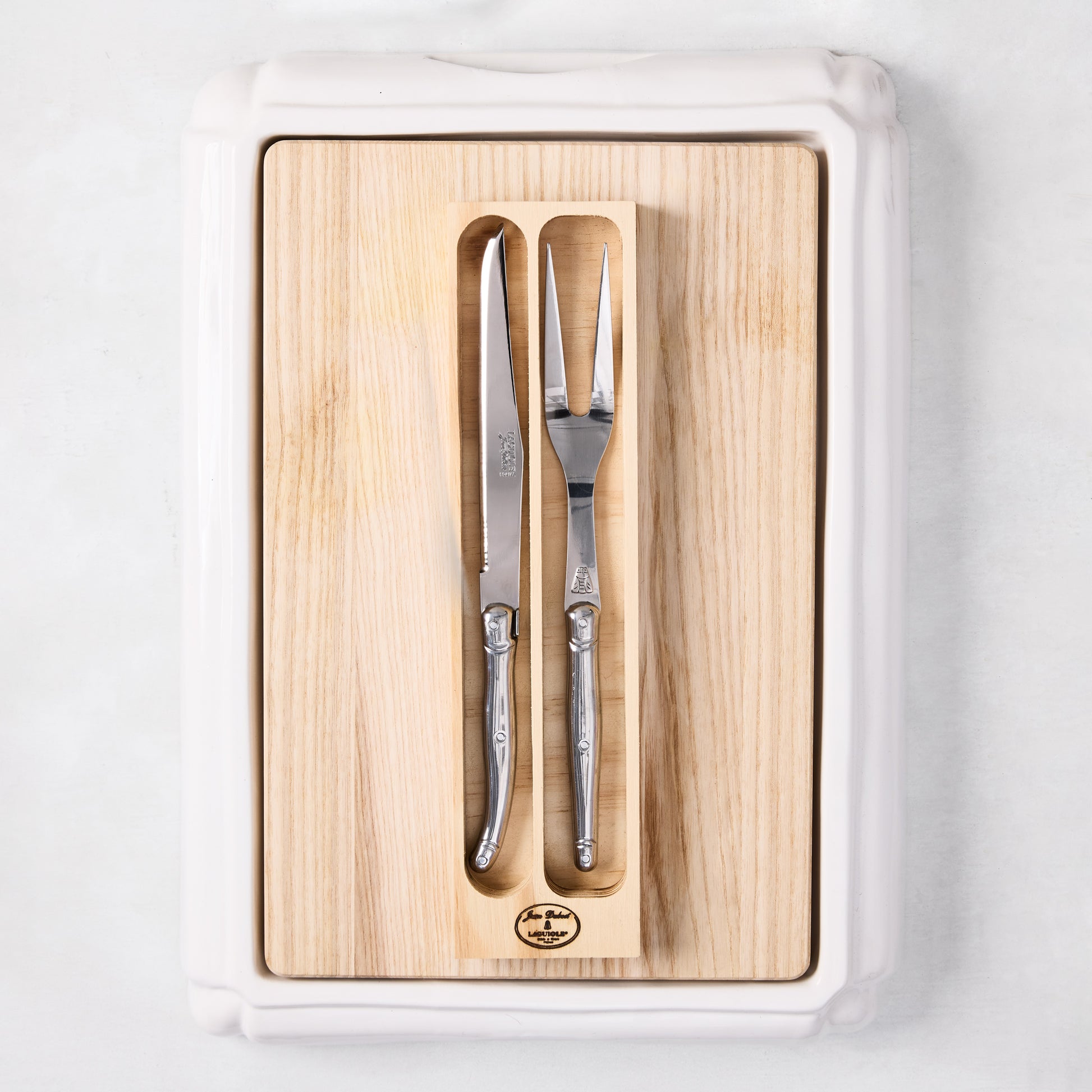 Jean Dubost stainless steel carving knife and fork set on a removable oak wood insert inside a white ceramic cutting board.