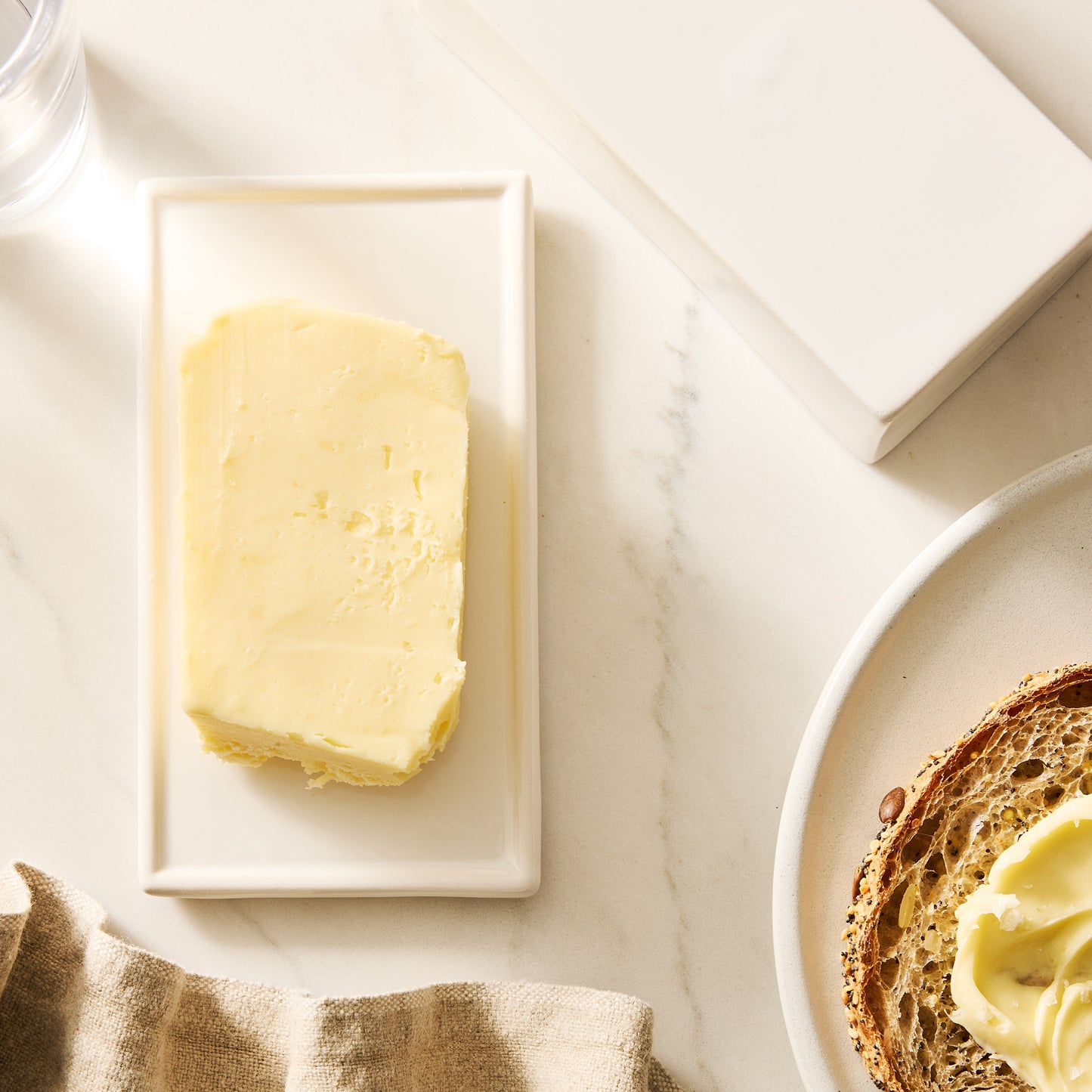 White ceramic butter dish with a stick of European butter, next to toast and a linen napkin.