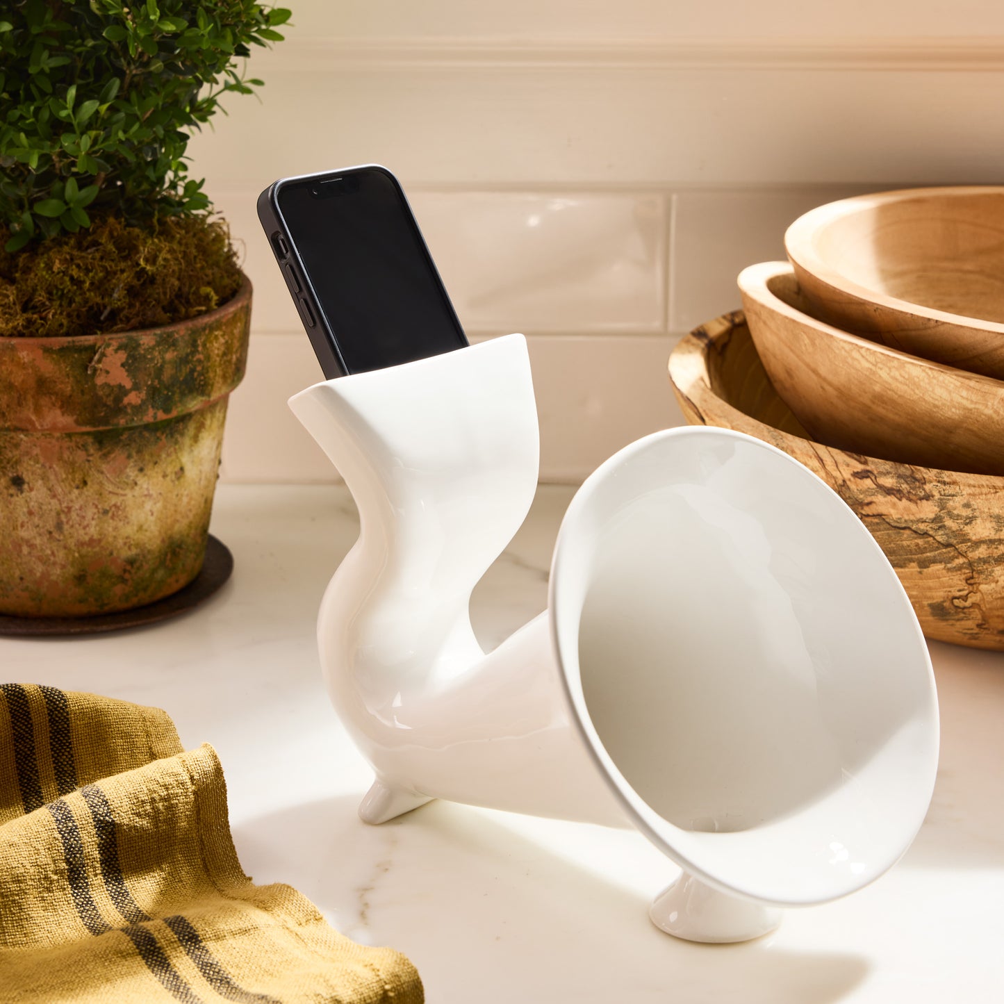 White ceramic kitchen speaker with a smartphone inserted, featuring a curved design and horn-shaped opening.