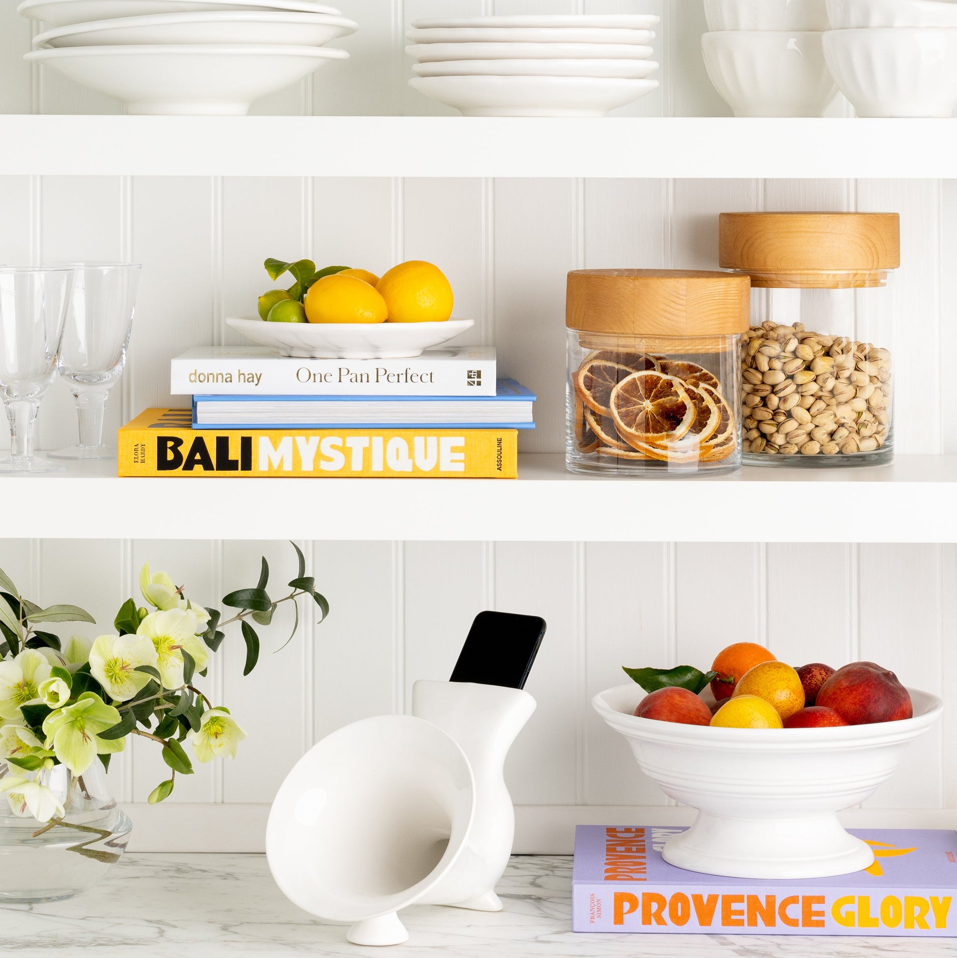 White ceramic kitchen speaker with a smartphone inserted, on a marble counter next to fruit and flowers.