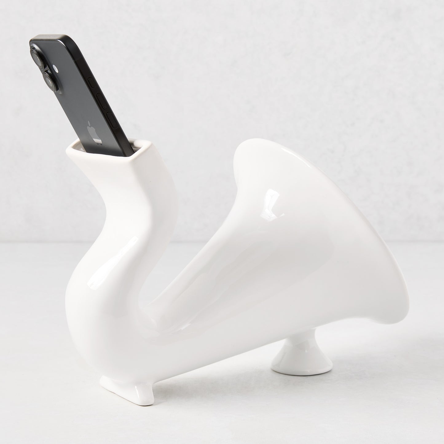 White ceramic speaker, 10"L, with a black smartphone inserted, featuring a curved, horn-like design.