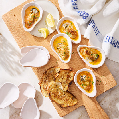 White ceramic oyster shells with baked oysters, lemon wedges, and toast on a wooden board.