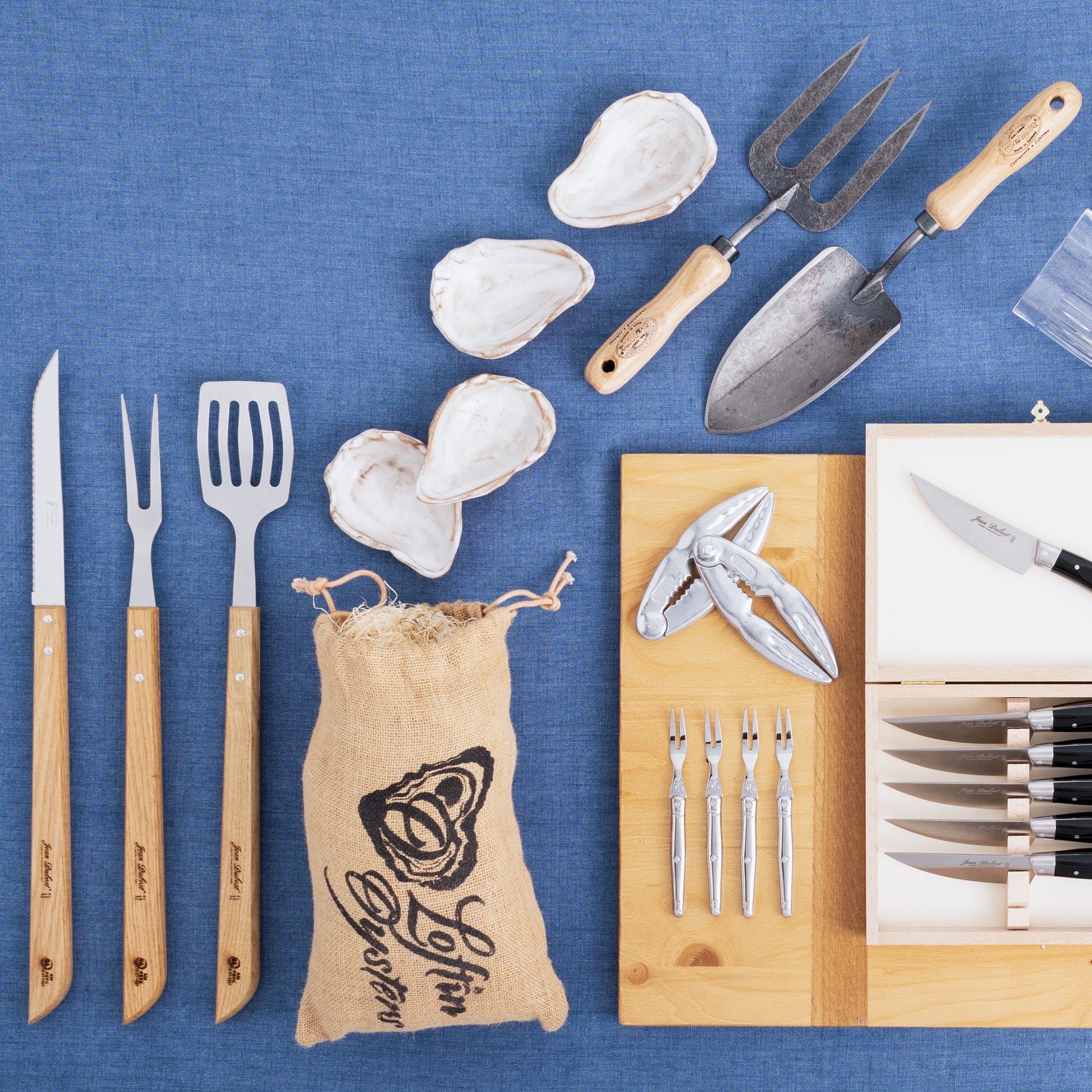White ceramic oyster shells, grilling tools with wood handles, oyster forks, and a cracker on blue fabric.