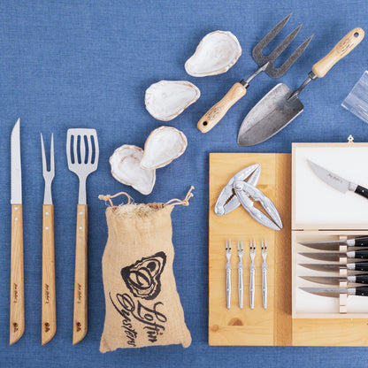White ceramic oyster shells, grilling tools with wood handles, oyster forks, and a cracker on blue fabric.