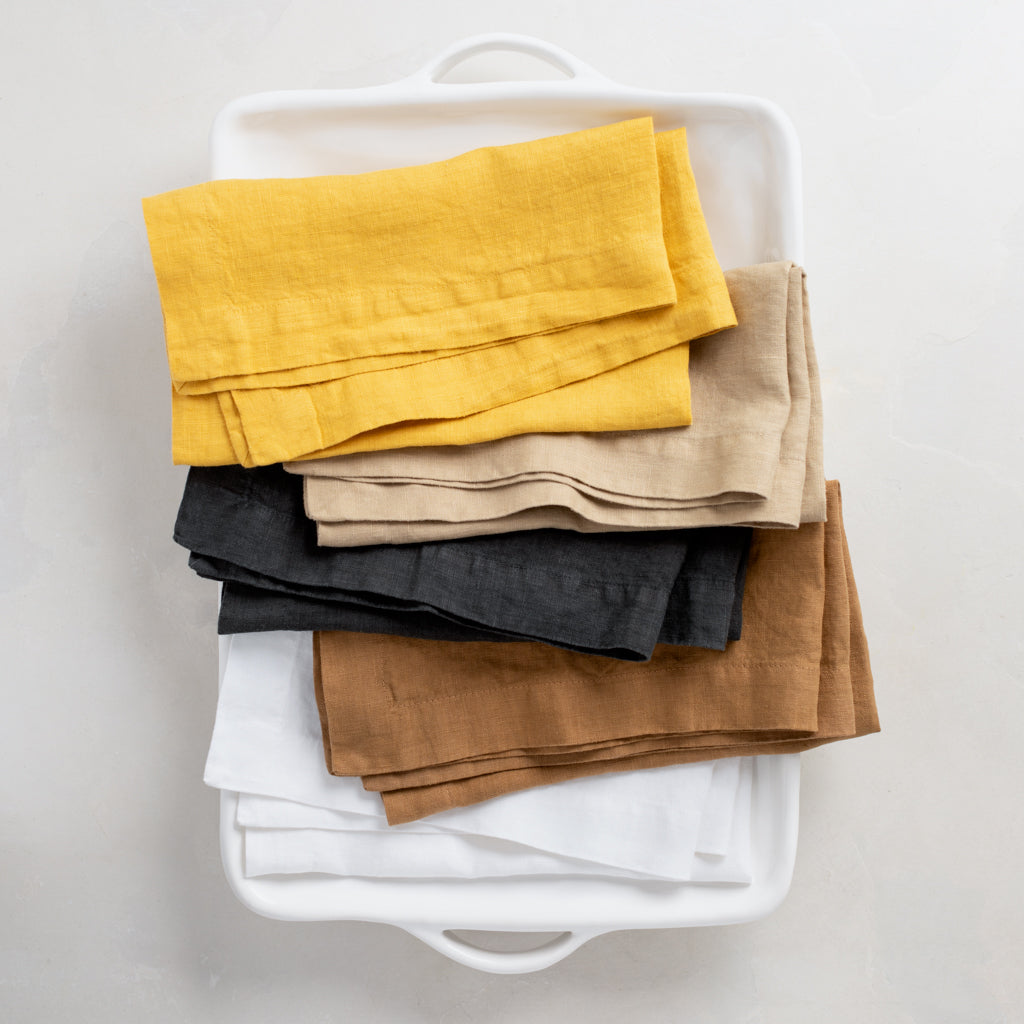 Stacked hand-dyed linen napkins in yellow, beige, charcoal, brown, and white in a white tray.