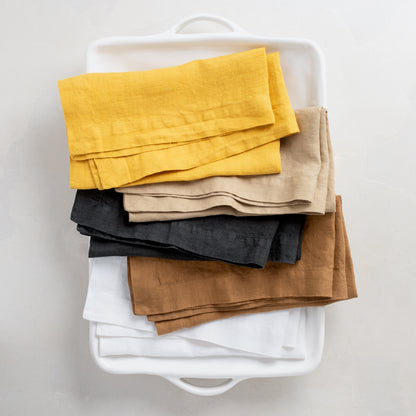 Stacked hand-dyed linen napkins in yellow, beige, charcoal, brown, and white in a white tray.