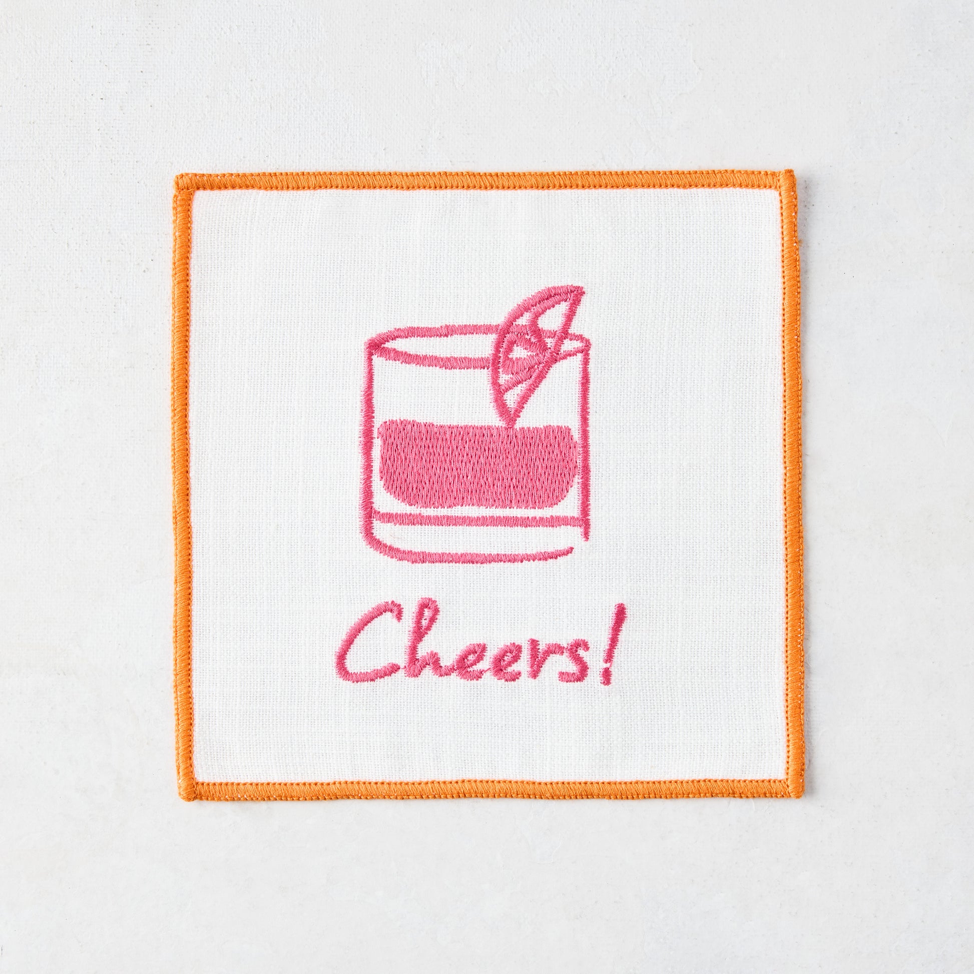 White cotton coaster with orange trim, pink embroidered cocktail and "Cheers!" text.