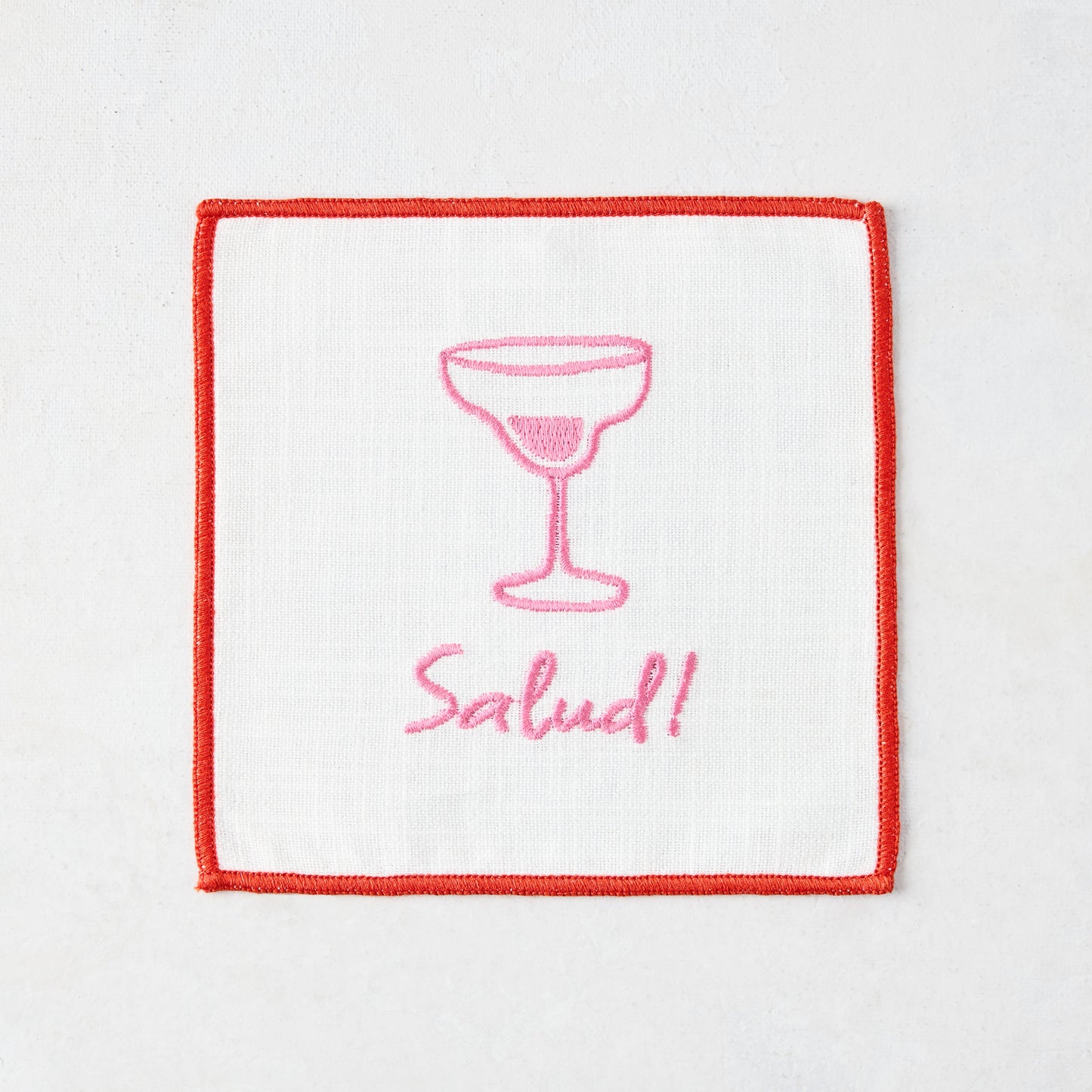 White cotton coaster with red trim, pink embroidered cocktail glass, and "Salud!" text.