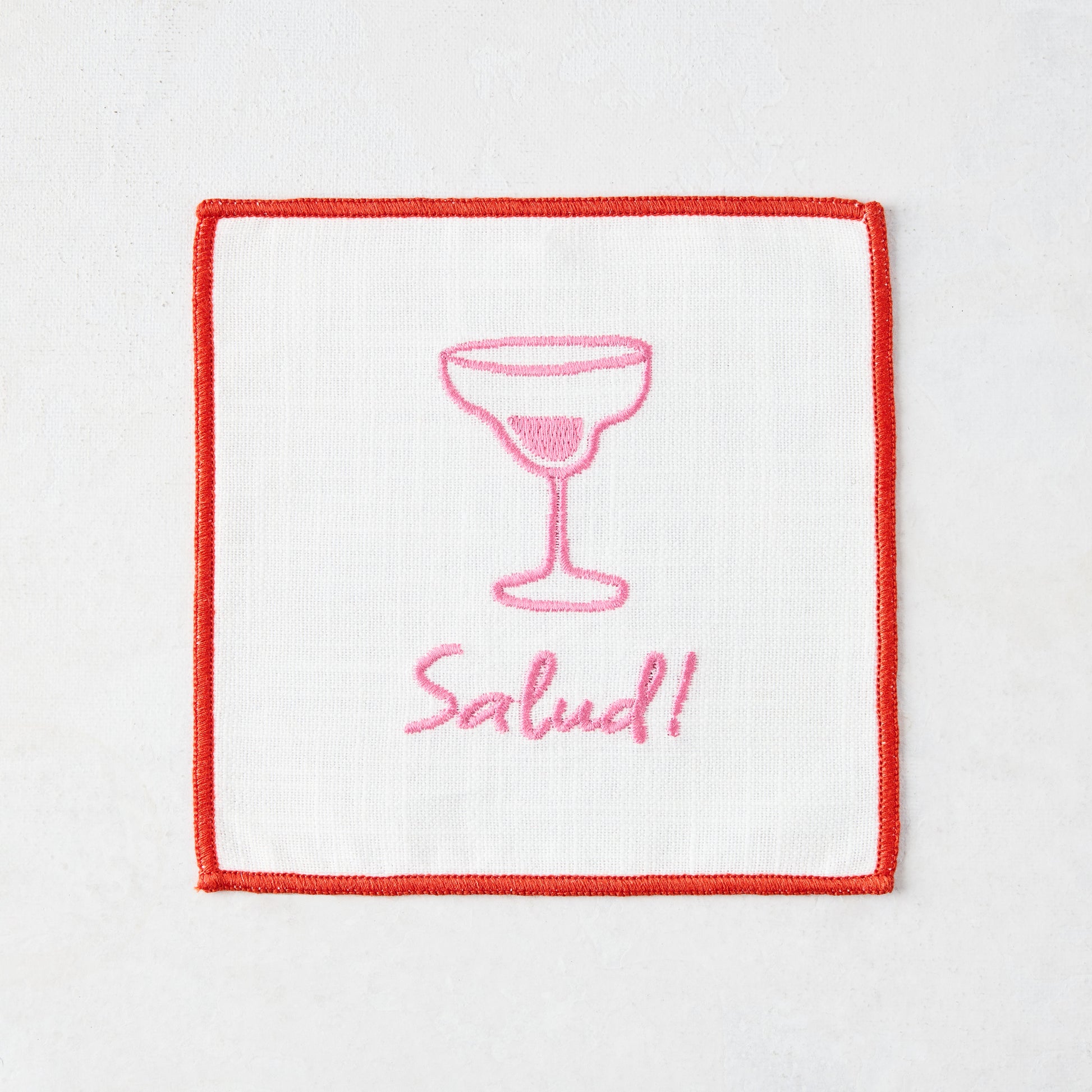 White cotton coaster with red trim, pink embroidered cocktail glass, and "Salud!" text.
