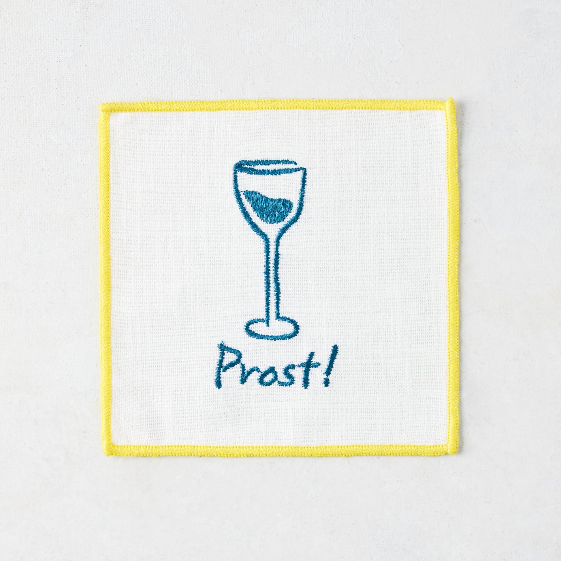 White cotton coaster with yellow trim, blue embroidered wine glass, and "Prost!" text.
