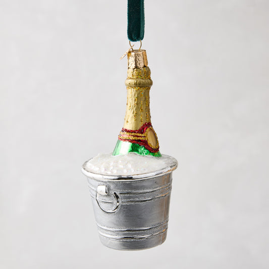 Hand-painted glass champagne bottle ornament in a silver bucket with white glitter foam, hanging from a green ribbon.
