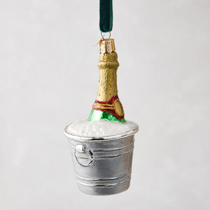 Hand-painted glass champagne bottle ornament in a silver bucket with white glitter foam, hanging from a green ribbon.