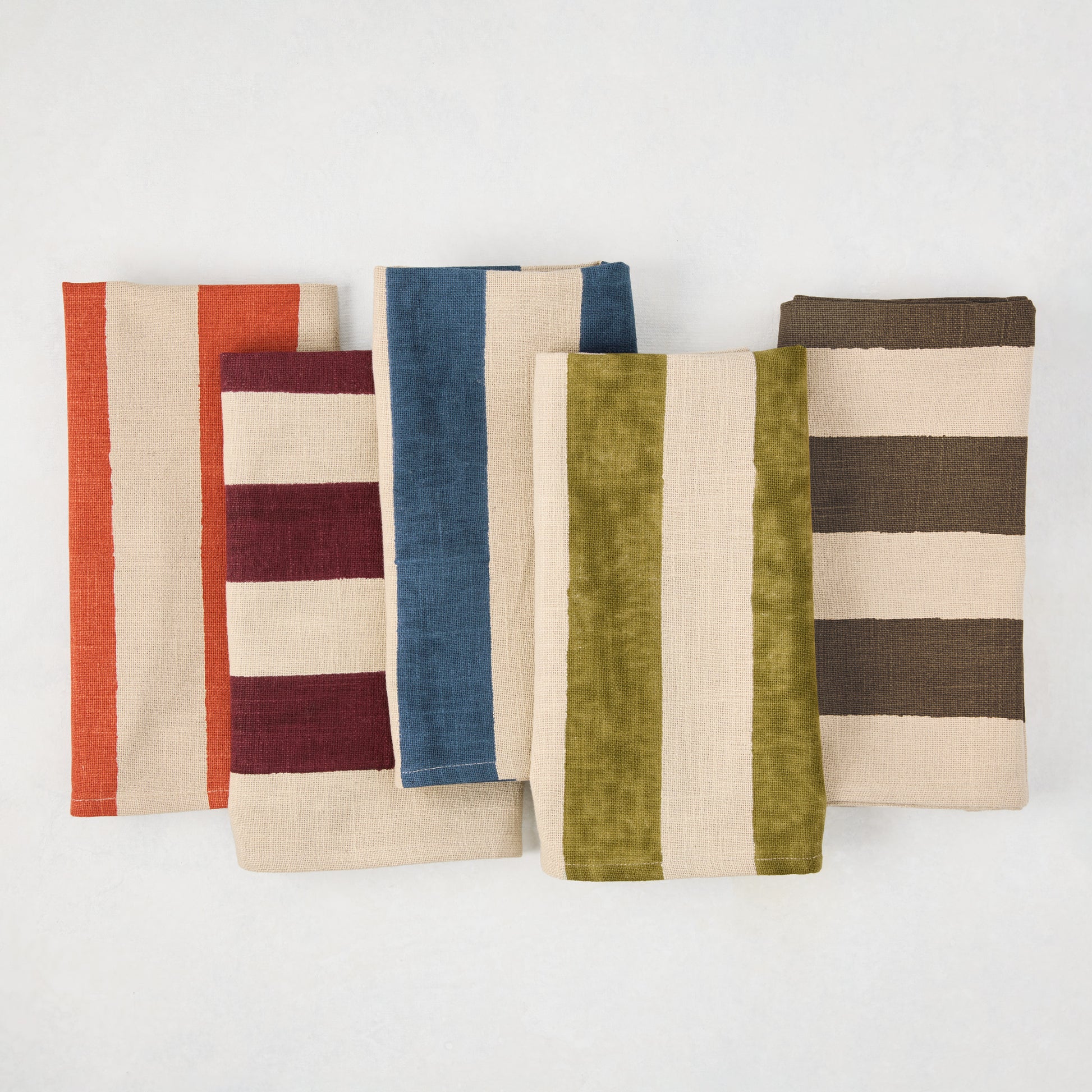 Five folded cotton napkins with wide vertical stripes in orange, red, blue, green, and chocolate brown on a khaki base.