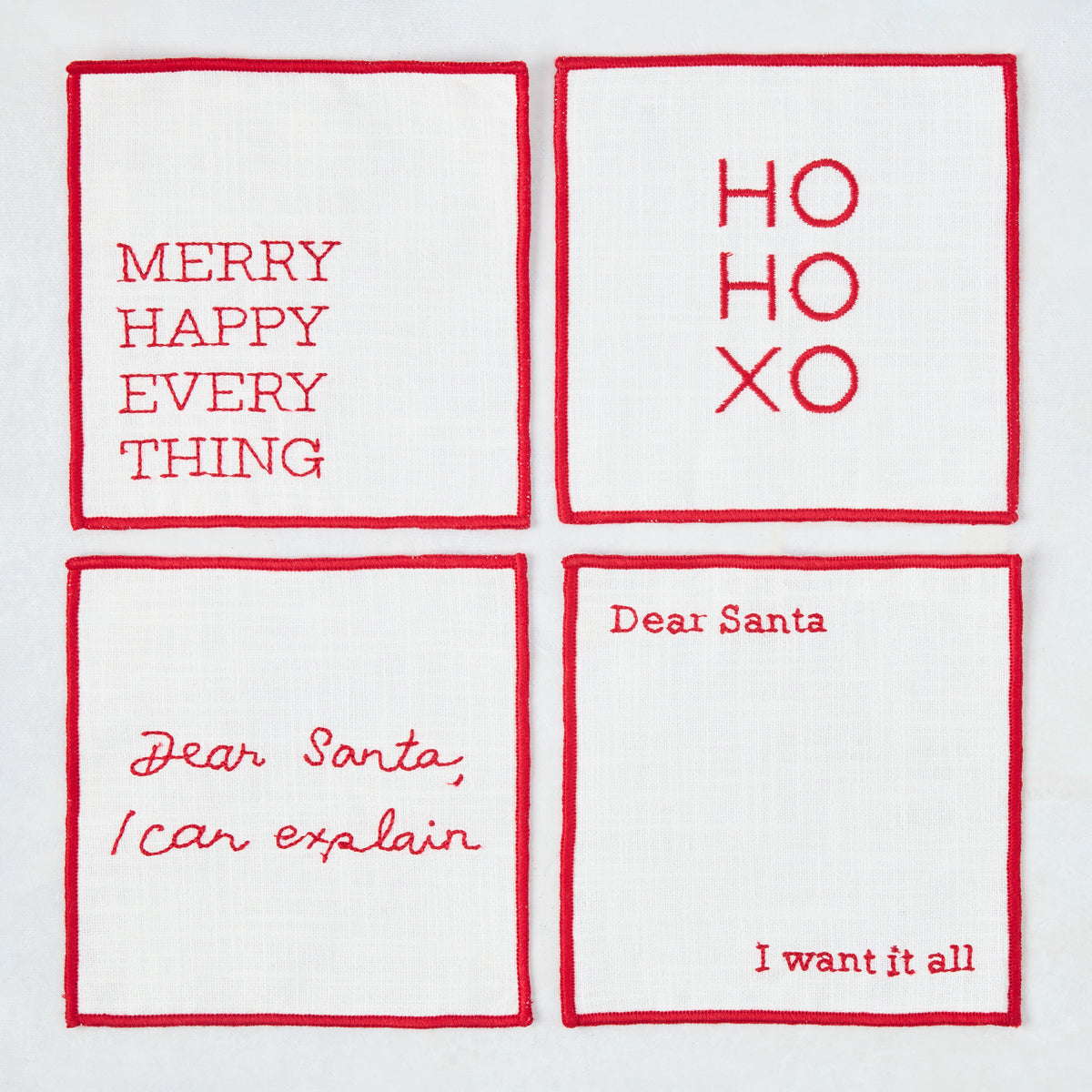 Set of four 5.5-inch square white cotton coasters with red embroidered Christmas phrases and red stitched borders.
