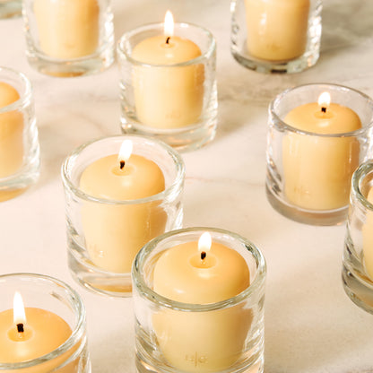 Clear glass votive candle holders with lit amber votive candles on a light surface.