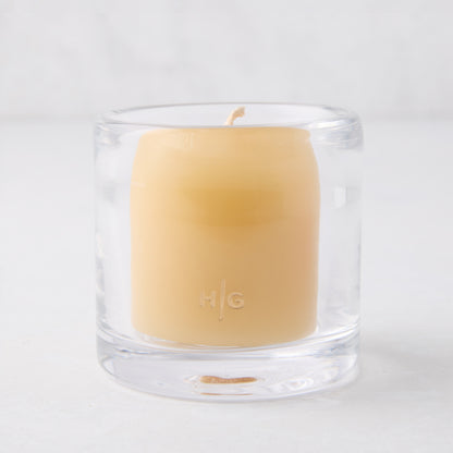 Clear glass votive candle holder with a light yellow candle inside, featuring an "HG" logo.
