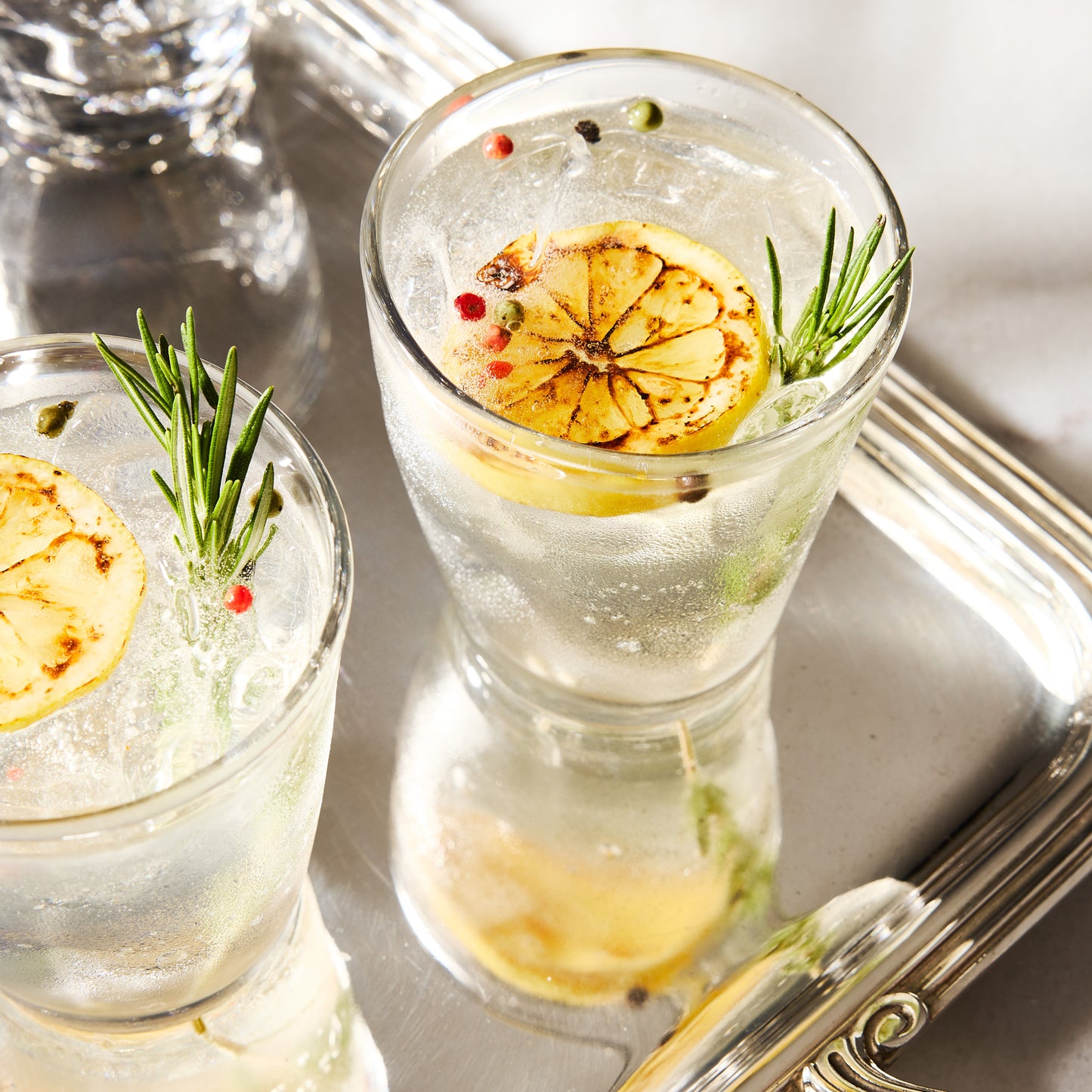 Two clear Henri 8 oz. glasses with drinks, garnished with grilled lemon and rosemary, on a silver tray.