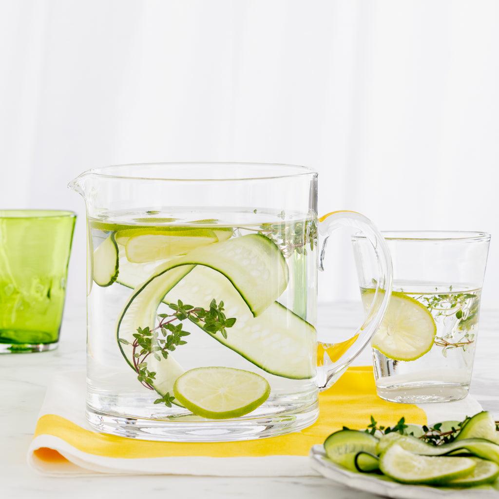 Clear Henri glass pitcher with cucumber, lime, and thyme infused water, next to a matching glass and green Henri glass.