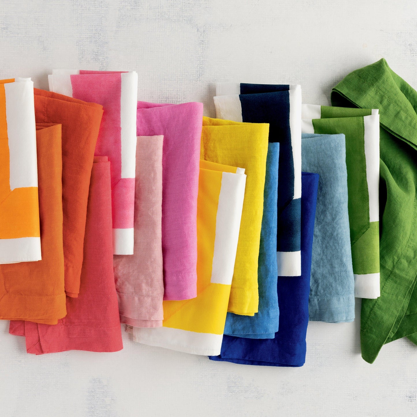 Row of colorful hand-dyed linen napkins in various shades, including Clover Pink, folded on a white surface.