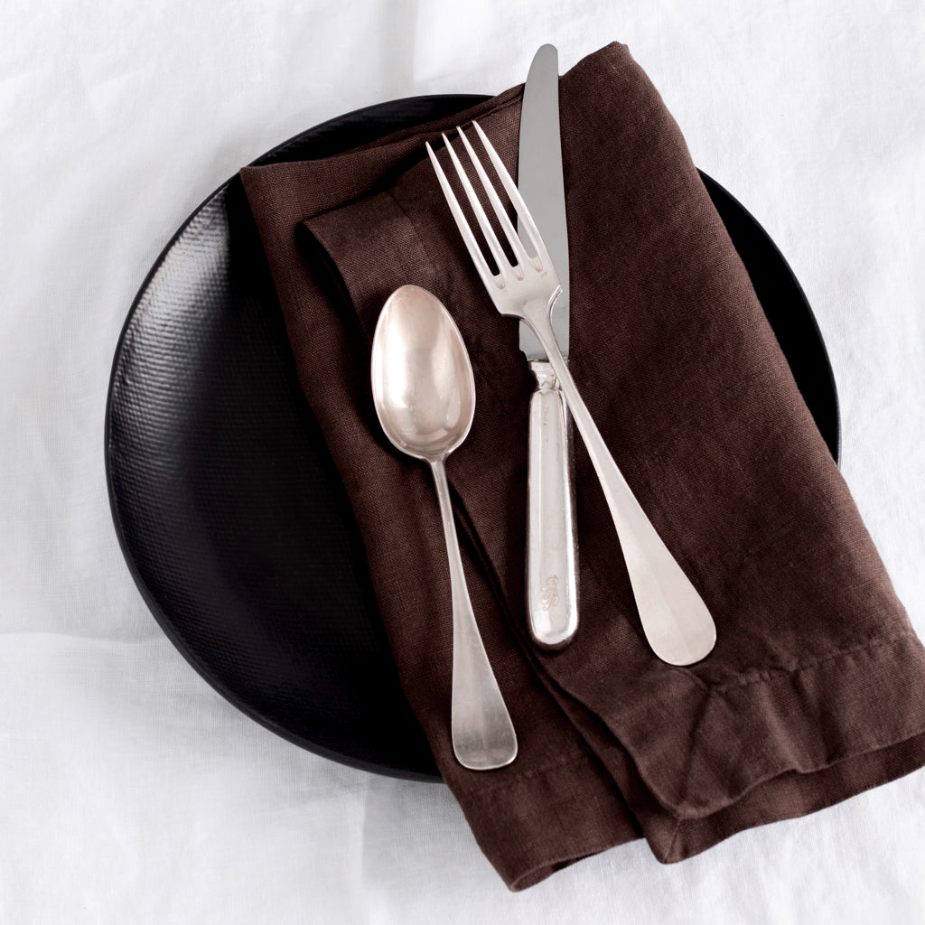 Cocoa HG brown hand-dyed linen napkin, 22", folded on a black plate with silver cutlery.