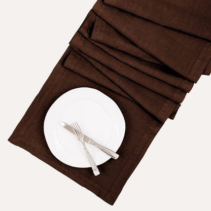 Cocoa HG hand-dyed linen table runner with a white plate and silverware, folded on a white background.