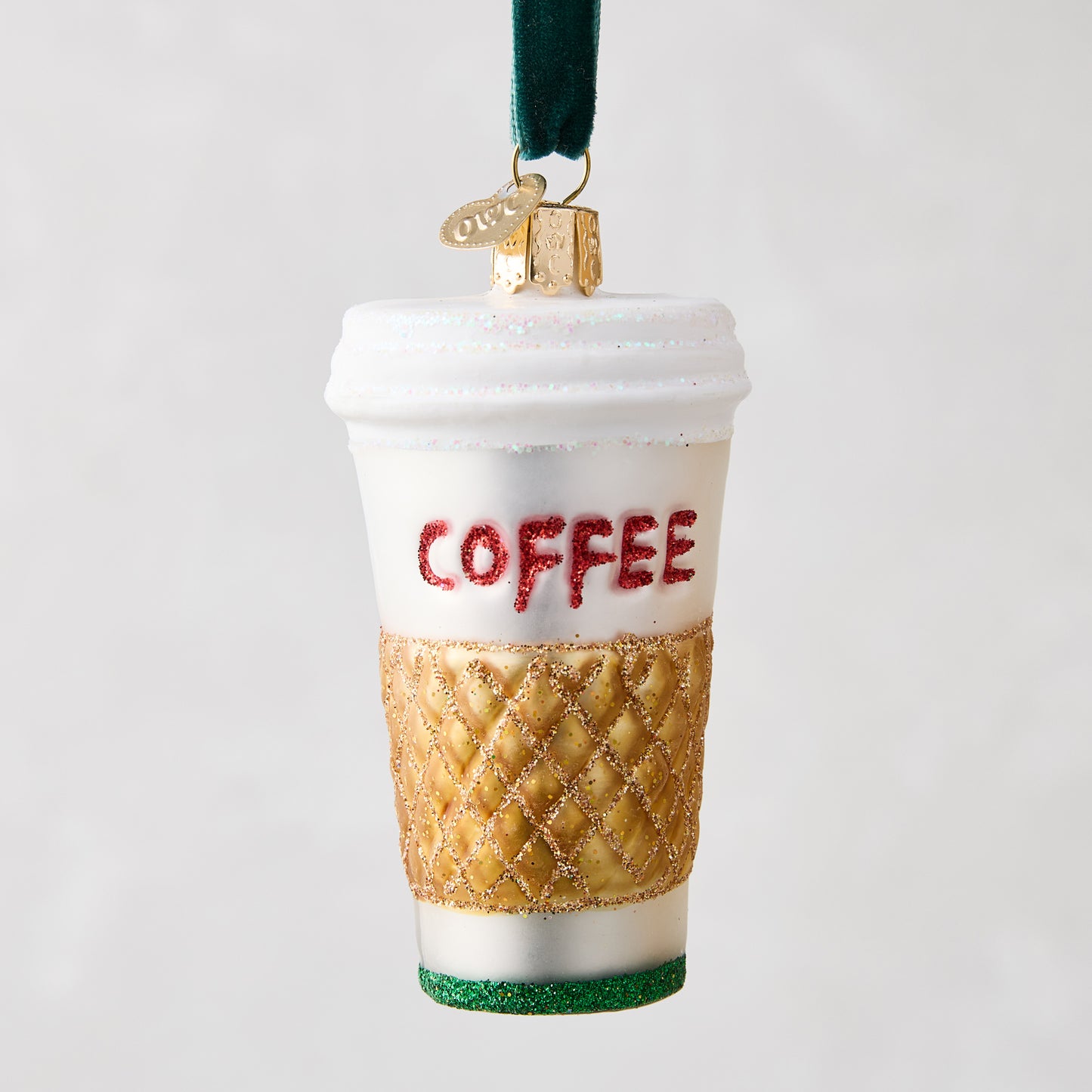 White glass coffee cup ornament with "COFFEE" in red glitter, gold quilted sleeve, and green glitter base.