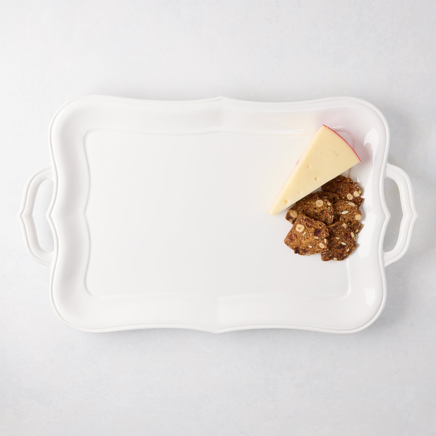 White ceramic Colette serving platter with scalloped edges and handles, holding cheese and crackers.