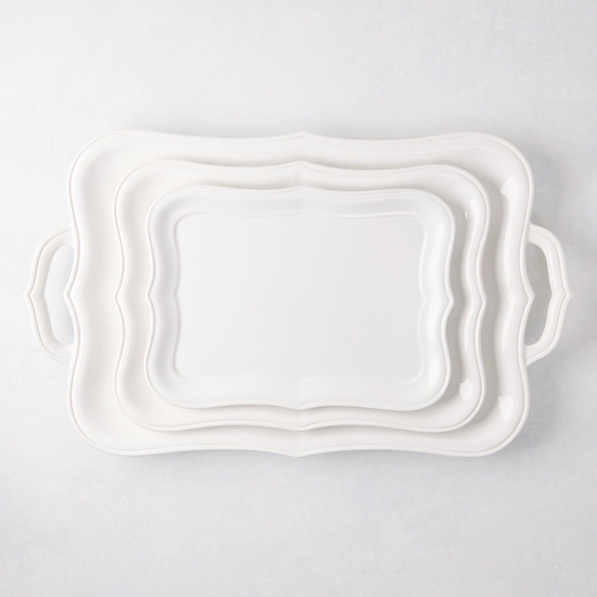 Three white Colette ceramic serving platters, scalloped edges, stacked, largest with handles.