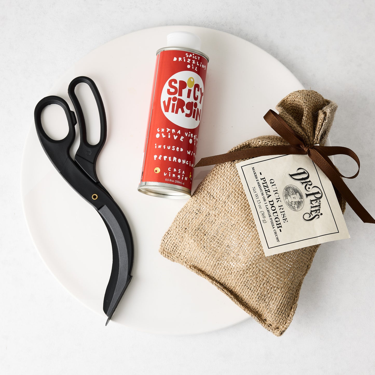 White ceramic pizza platter with black pizza scissors, a red bottle of spicy olive oil, and a burlap bag of pizza dough.