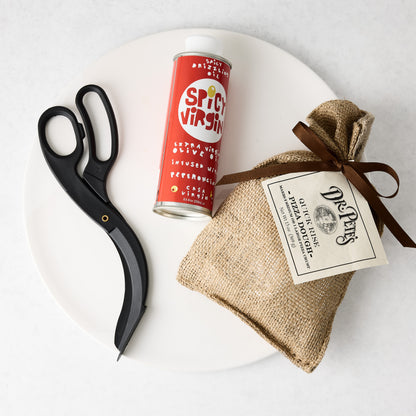 White ceramic pizza platter with black pizza scissors, a red bottle of spicy olive oil, and a burlap bag of pizza dough.