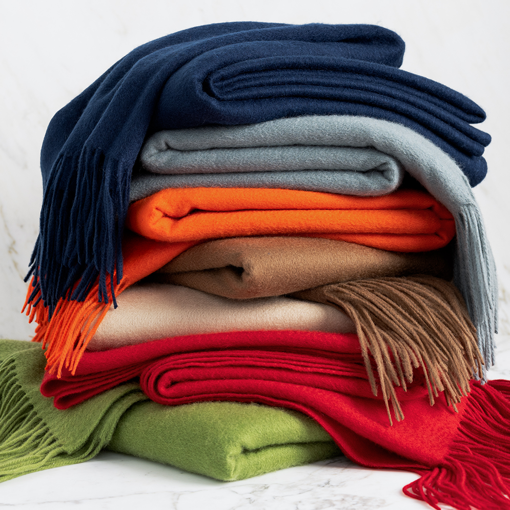 Stack of folded cashmere throw blankets in navy, grey, orange, tan, cream, red, and lime green with fringe.