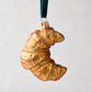 Hand-painted, glittered glass croissant ornament with a gold cap and green velvet ribbon.