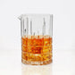 Clear square-cut crystal cocktail mixing glass with a pour spout, filled with amber liquid and ice.