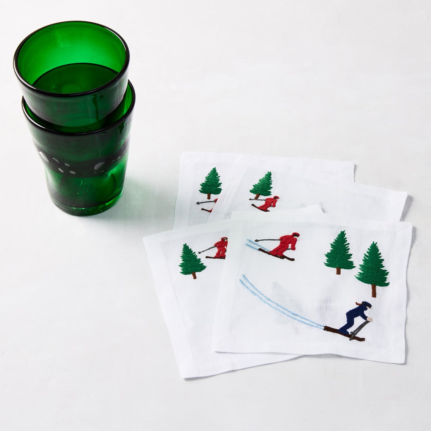 Two dark green Henri mouth-blown glass tumblers next to white cocktail napkins with embroidered skiers and pine trees.