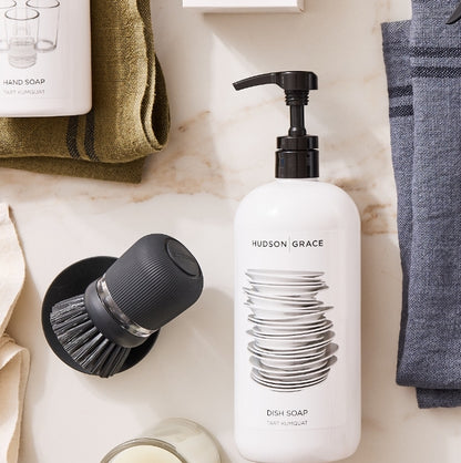 Dark grey soap dispensing dish brush with stand, next to a white dish soap bottle.