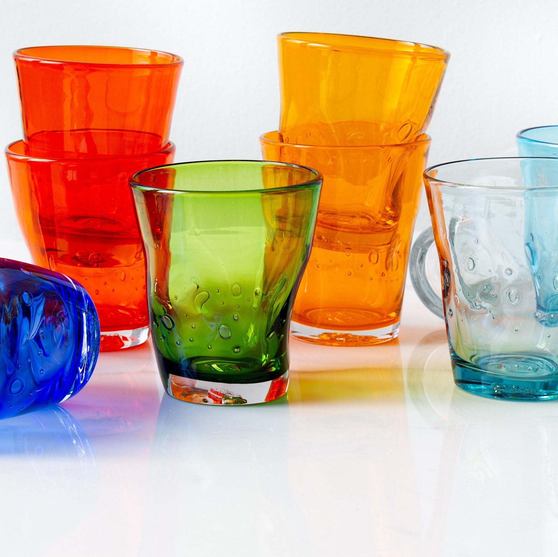 Assorted Henri mouth-blown glasses in dark orange, green, blue, and clear with unique bubbles and asymmetry.
