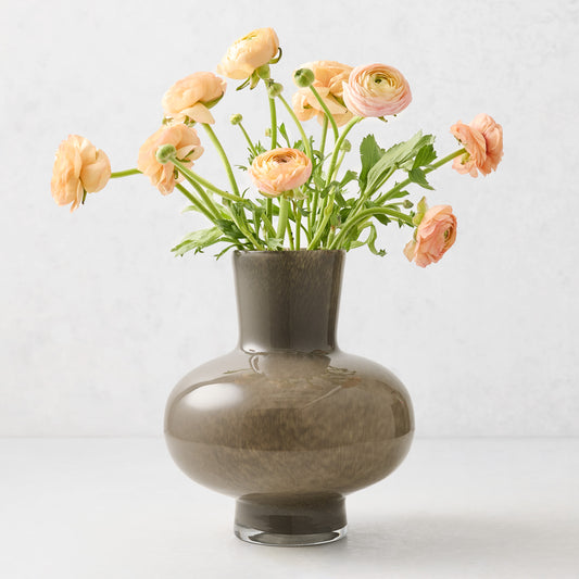 Hand-blown dark taupe glass vase with a geometric silhouette and curved edges, filled with peach ranunculus flowers.