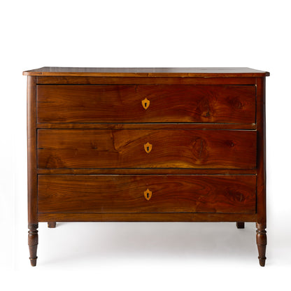 Dark wood dresser with three drawers, mahogany veneer, brass keyholes, and turned feet in a French empire style.