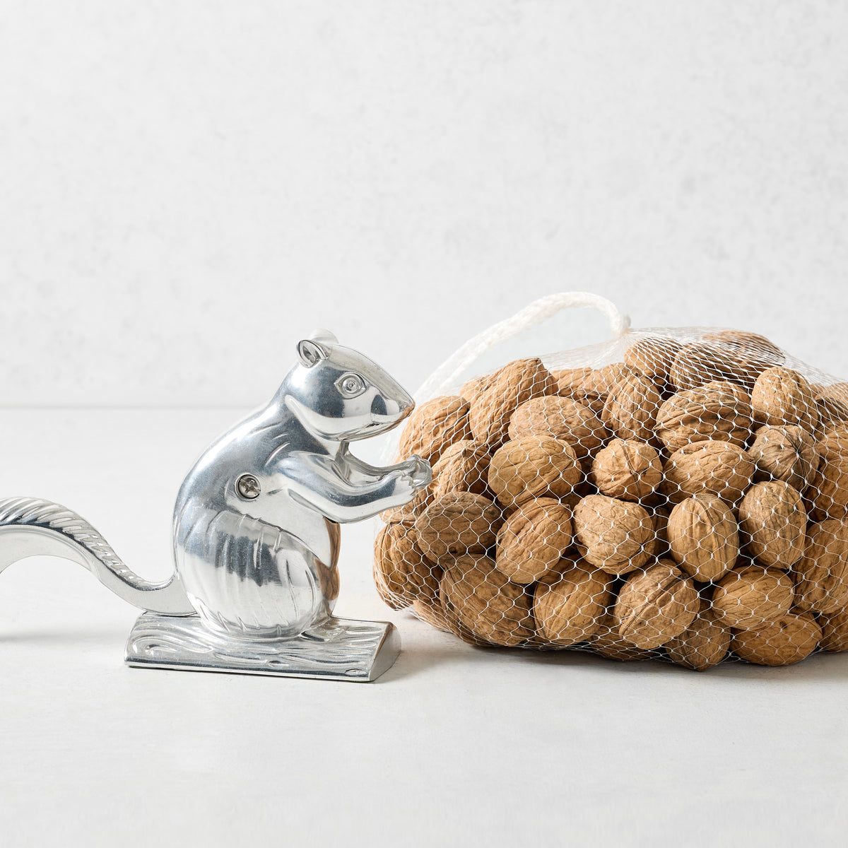 Silver squirrel-shaped metal nutcracker next to a mesh bag of whole walnuts on a white surface.