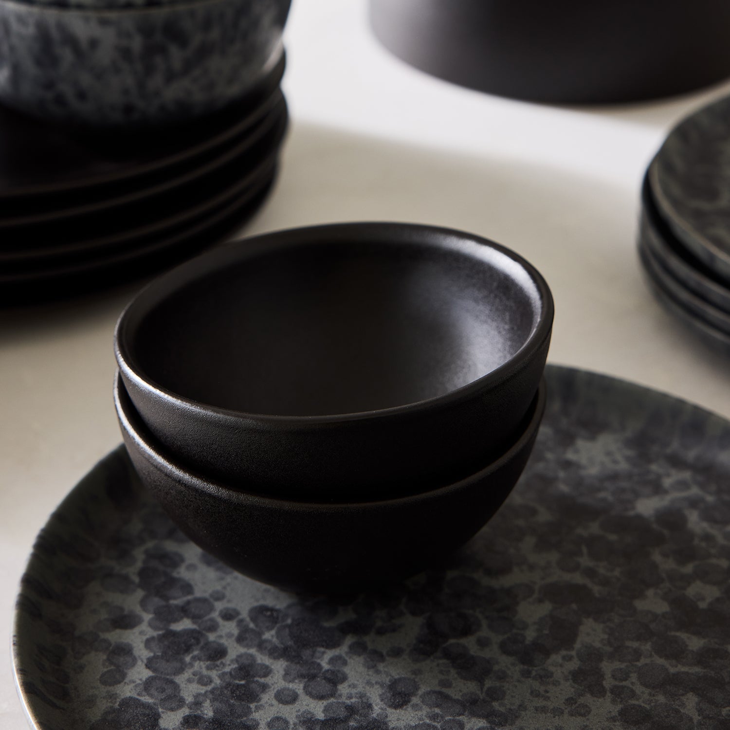 Two stacked Diane Keaton + Hudson Grace matte black graphite stoneware individual bowls on a speckled plate.