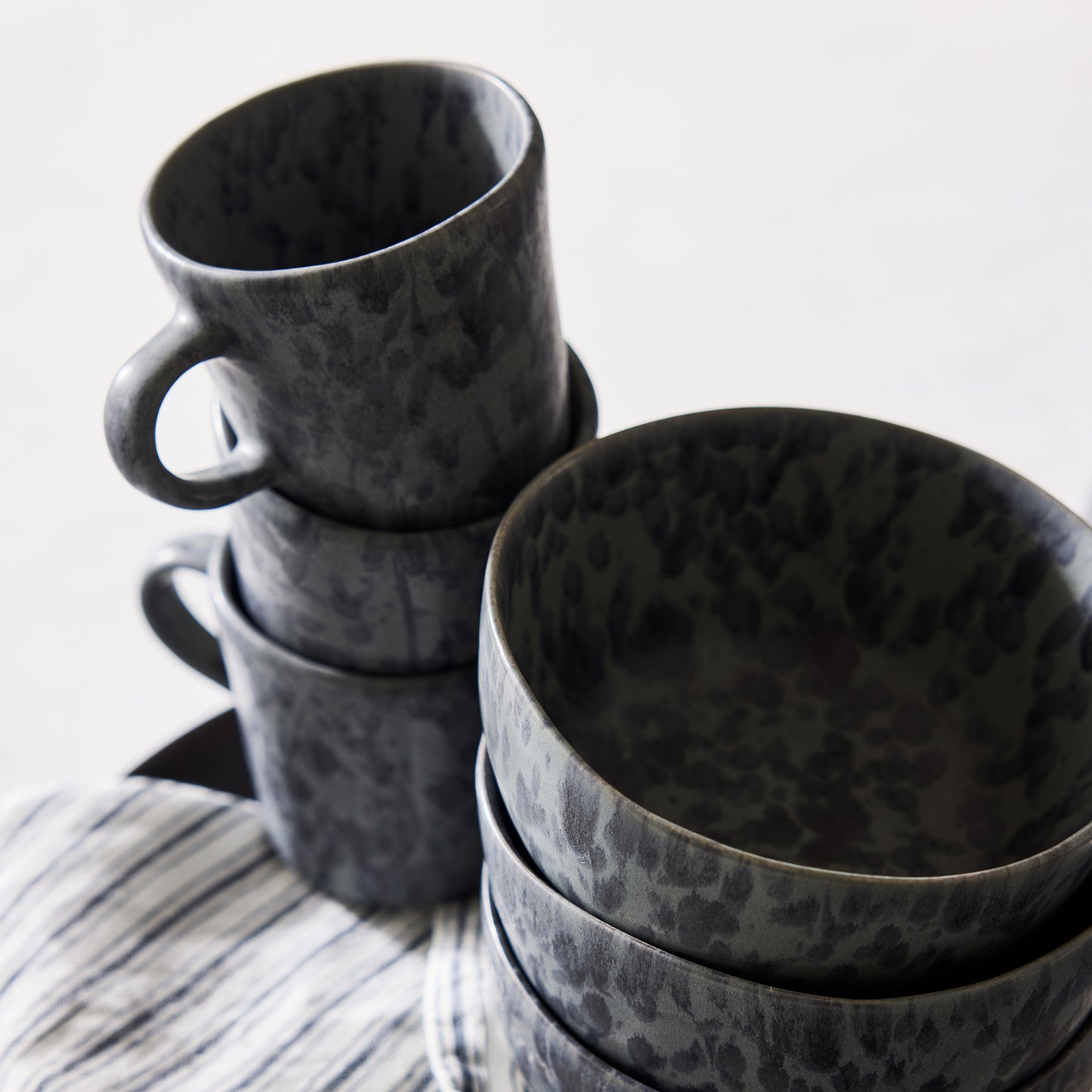 Diane Keaton + Hudson Grace black and grey splatter stoneware mugs and bowls, stacked on a striped cloth.