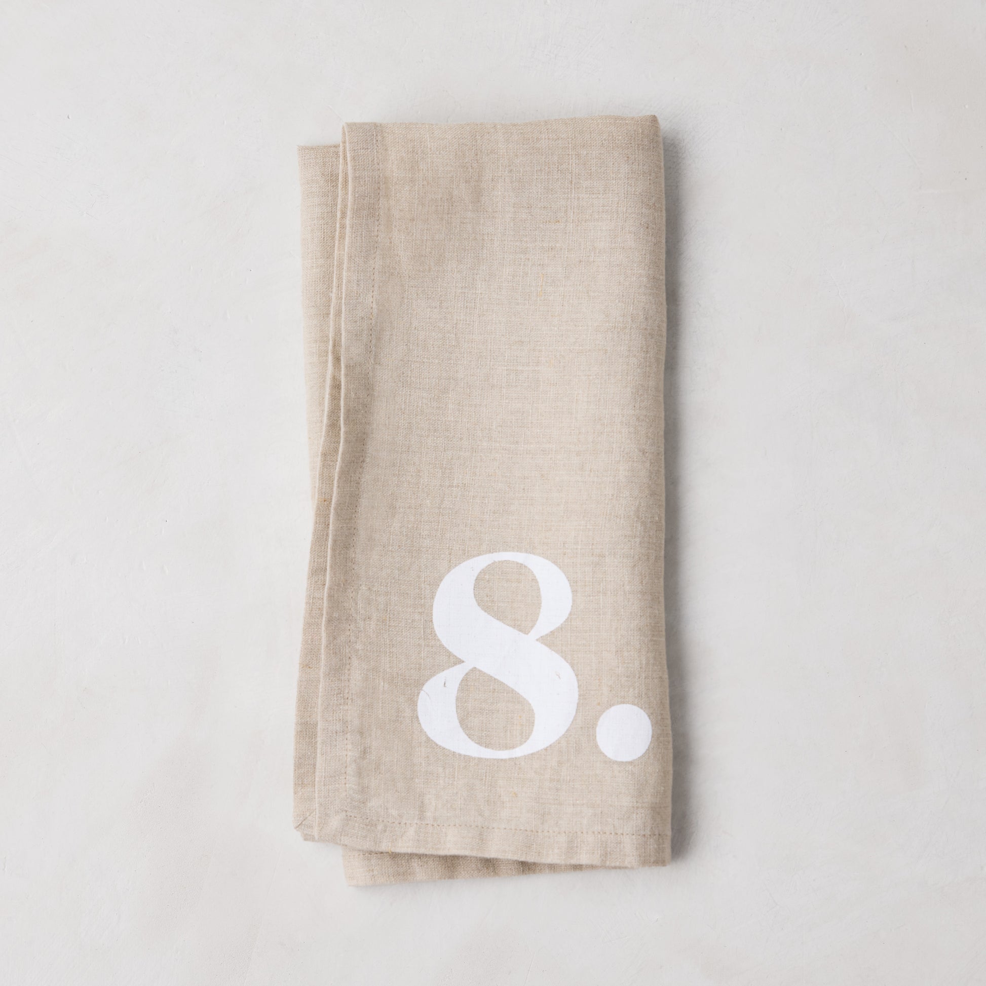 Diane Keaton + Hudson Grace Khaki Linen Numeric Napkin, folded, with a white number 8.